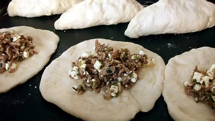 Rohe B&auml;rlauch-Pogaca mit Hackfleisch und Feta-F&uuml;llung auf einem Backblech, geformte Teigtaschen warten auf das Backen