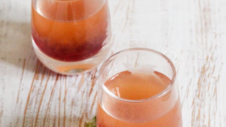 Zwei Gl&auml;ser mit einem rosafarbenen Getr&auml;nk stehen auf einem hellen Holztisch, umgeben von einer Erdbeere und Minze.