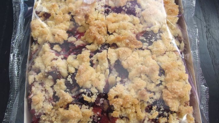 Ein tiefgek&uuml;hlter Kuchen mit knusprigem Belag liegt in einer transparenten Verpackung auf einem dunklen Tisch.