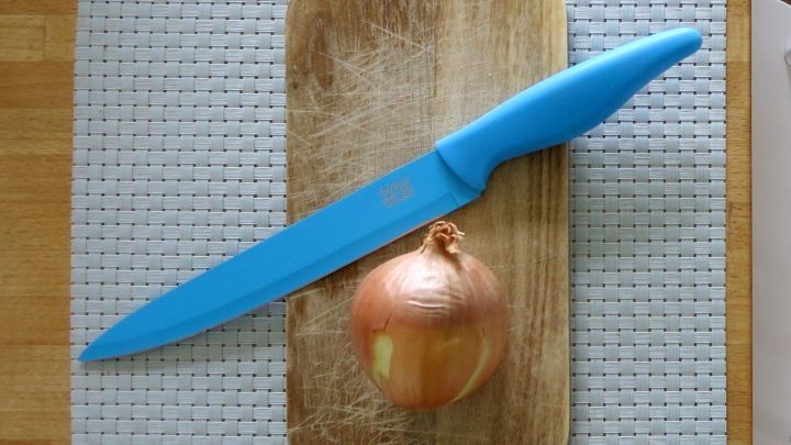 Eine Zwiebel liegt auf einem Holzbrett, w&auml;hrend ein t&uuml;rkiser Messer daneben bereit zum Schneiden ist.