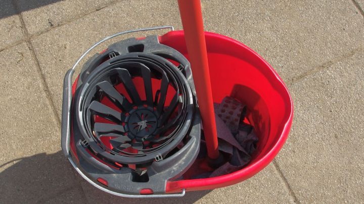 Ein roter Eimer mit einem Wischmopp und einem ausgeklappten Wischkopf steht auf einem Gehweg im Freien.