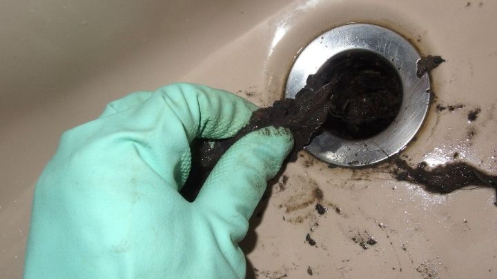 Eine Hand mit einem gr&uuml;nen Handschuh zieht Schmutz aus einem Abfluss, der schmutzig aussieht und gepflegt werden sollte.
