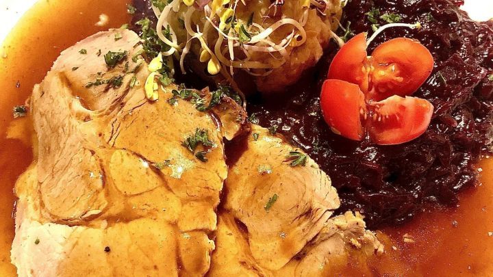 Saftiger Schweinebraten liegt auf einem Teller, umgeben von Rotkraut, Kr&auml;utern und Tomaten im gem&uuml;tlichen Restaurant.