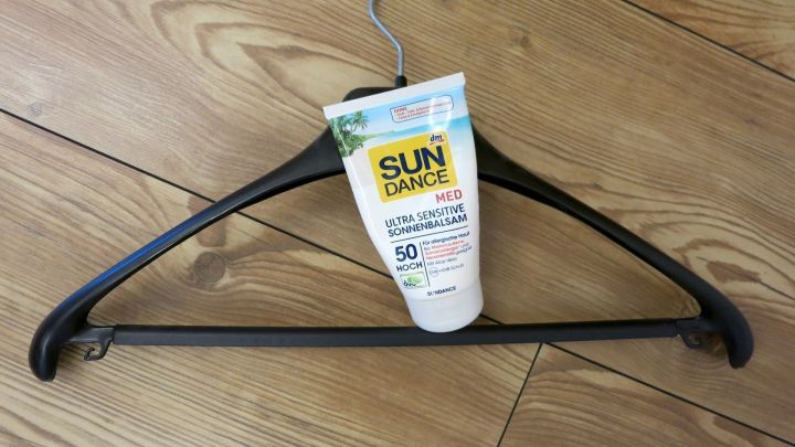 Kleiderb&uuml;gel h&auml;lt Sonnencreme "SUN DANCE MED Ultra Sensitive Sonnenbalsam 50 Hoch" auf Holzfu&szlig;boden.