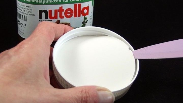 Eine Hand h&auml;lt einen ge&ouml;ffneten Nutella-Deckel, w&auml;hrend ein L&ouml;ffel bereit ist, um die Creme zu entnehmen.
