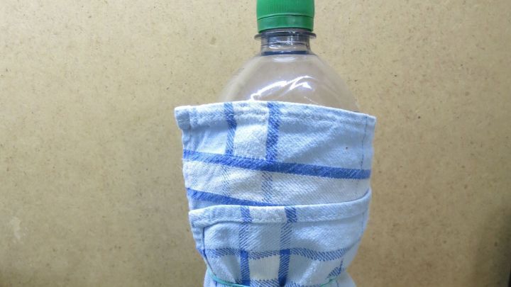 Eine Plastikflasche ist eng in ein blau-wei&szlig; kariertes K&uuml;chentuch gewickelt, steht vor einem neutralen Hintergrund.