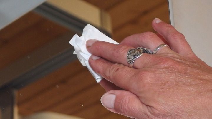 Eine Hand mit einem Brillenputztuch reinigt einen Spiegel in einem hellen, warmen Raum mit Holzboden.