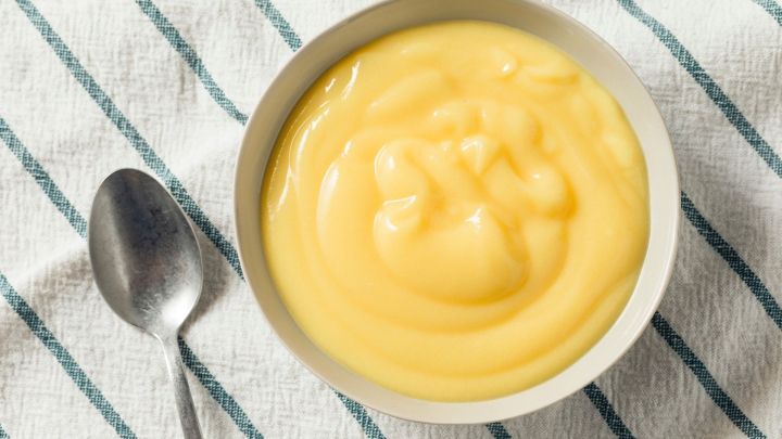 Eine Sch&uuml;ssel mit cremigem Pudding steht auf einem gestreiften Tuch, bereit zum Genie&szlig;en, neben einem L&ouml;ffel.