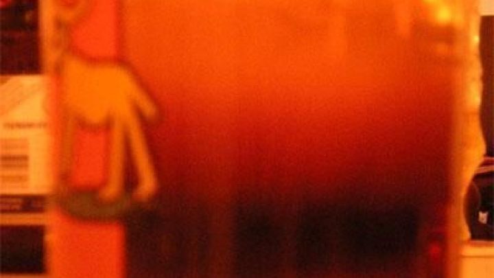 Ein Glas mit einem mehrschichtigen Getr&auml;nk steht auf einem Tisch, umgeben von unscharfem, warmem Licht.