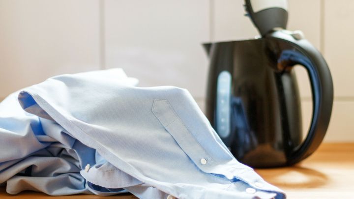 Ein Wasserkocher steht neben einem leicht zerknitterten, blauen Hemd auf einer Holzoberfl&auml;che.