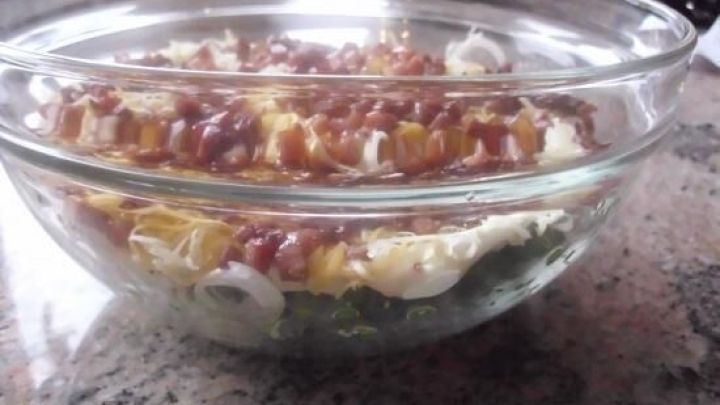 Eine Glassch&uuml;ssel zeigt einen Schichtsalat auf einem Granittisch, bestehend aus verschiedenen gr&uuml;nen, wei&szlig;en und braunen Zutaten.