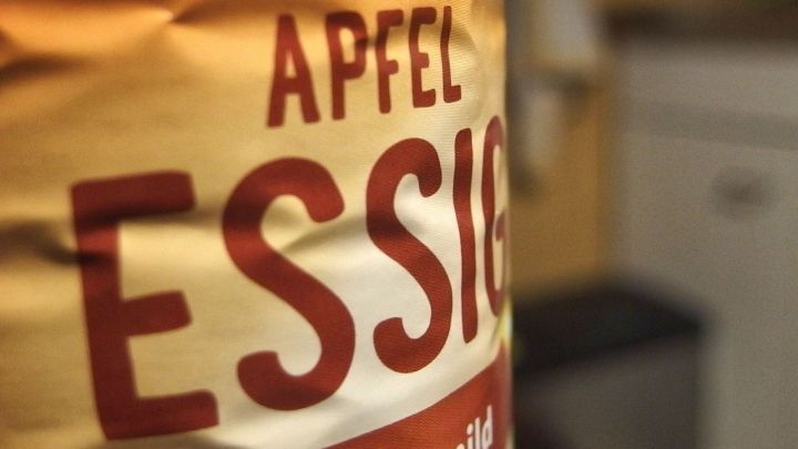 Eine Flasche Apfelessig steht im Vordergrund, w&auml;hrend der Hintergrund unscharf eine K&uuml;che zeigt.
