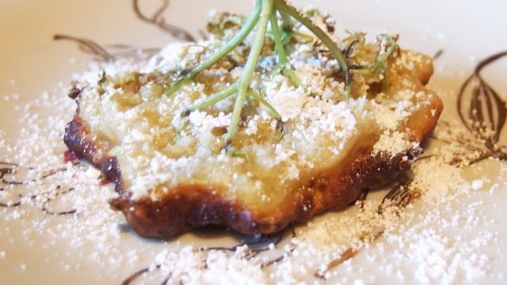 Ein gebackenes Holunderbl&uuml;tengeb&auml;ck liegt auf einem dekorativen Teller, best&auml;ubt mit Puderzucker und einem Hauch von Kr&auml;utern.