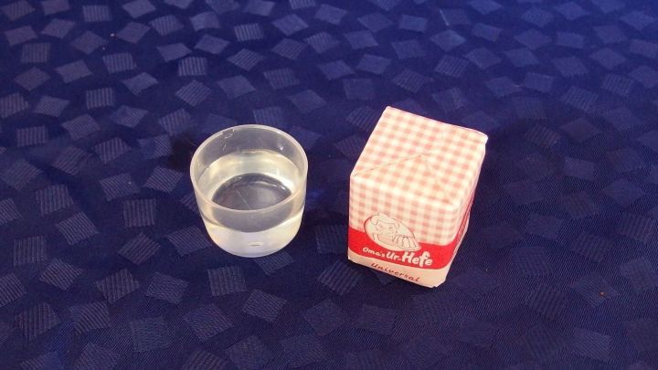 Eine klare Sch&uuml;ssel mit Wasser steht neben einer rot-wei&szlig;en Verpackung f&uuml;r Hefe auf einem blauen Tisch.