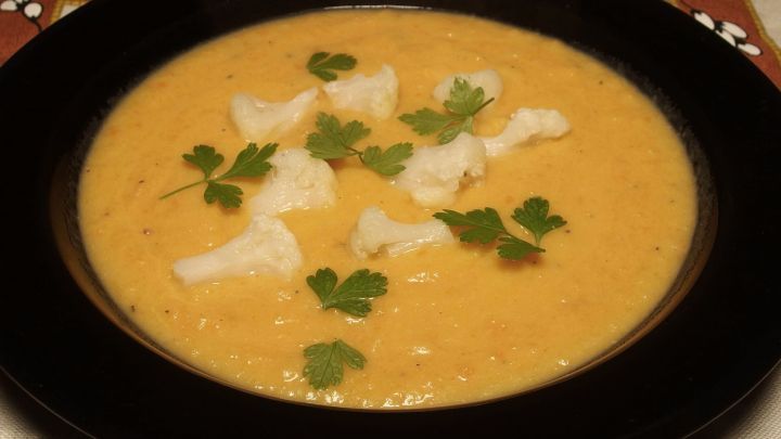 Eine cremige Blumenkohlcremesuppe wird in einer schwarzen Sch&uuml;ssel serviert, garniert mit Blumenkohlst&uuml;cken und frischem Gr&uuml;n.