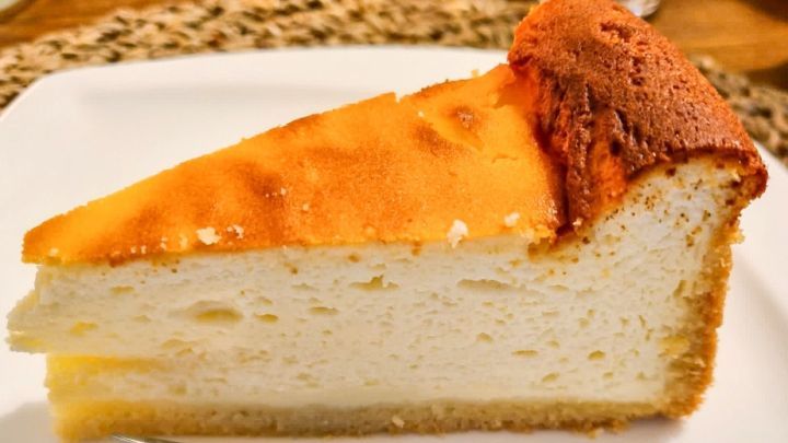 Ein St&uuml;ck Quarktorte liegt auf einem wei&szlig;en Teller, w&auml;hrend eine Gabel daneben bereitliegt, um es zu genie&szlig;en.