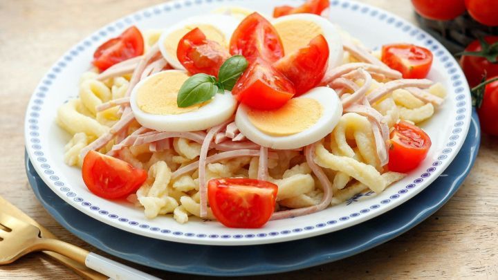 Ein Teller Sp&auml;tzle-Salat zeigt hellgelbe Sp&auml;tzle, Scheiben von hartgekochten Eiern und frische Tomaten, garniert mit Basilikum.