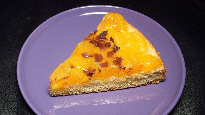 Ein Stück Obstkuchen liegt auf einem lila Teller, frisch angeschnitten und präsentiert sich in einer appetitlichen Gelb-Orange-Farbe.