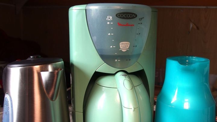 Eine gr&uuml;ne Kaffeemaschine steht in der Mitte zwischen einem silbernen Kessel und einer blauen Kanne auf einer K&uuml;che.