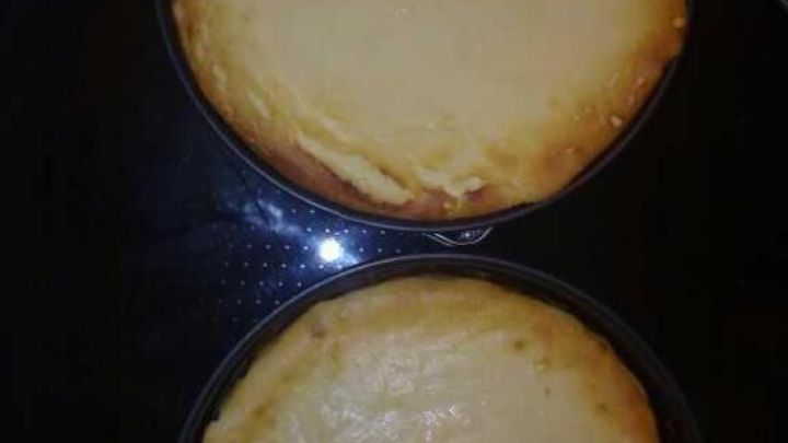 Zwei K&auml;sekuchen ohne Boden liegen in runden Formen auf einem dunklen Kochfeld, frisch aus dem Ofen.