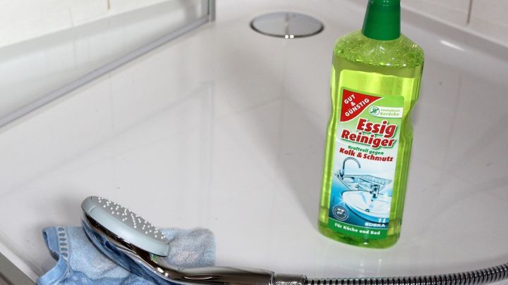 In einer Dusche steht eine Flasche Essigreiniger neben einem Reinigungstuch und einer Duschbrause auf dem fliesenboden.