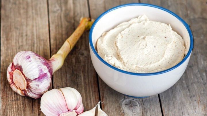 Eine Sch&uuml;ssel mit Knoblauch-Creme-Dip liegt auf einem Holztisch, umgeben von frischem Knoblauch.