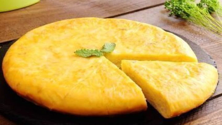 Eine goldbraune Tortilla de Patata liegt auf einem Schieferbrett mit einem herausgeschnittenen St&uuml;ck; Petersilie im Hintergrund.