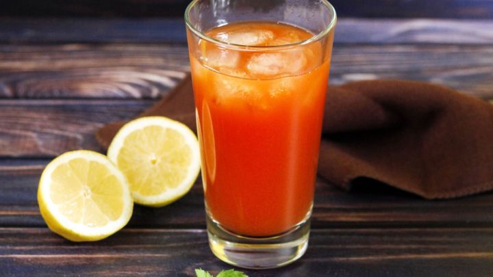 Ein Glas Tomatensaft mit Eisw&uuml;rfeln steht auf einem Holztisch, umgeben von zwei Zitronenh&auml;lften und einem braunen Tuch.