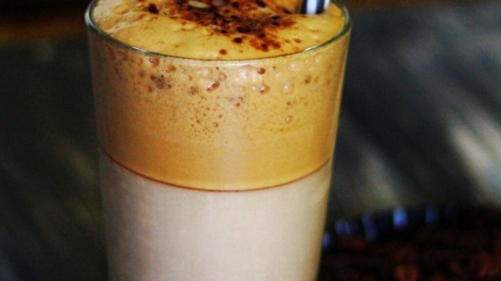 Ein hoher Eiskaffee mit Schaum und Kakao best&auml;ubt, steht auf einem Tisch mit einem gestreiften Strohhalm darin.