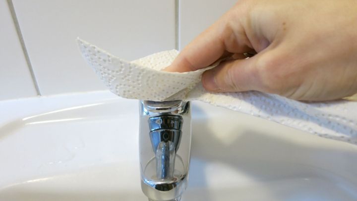Eine Hand h&auml;lt ein Papiertuch, um Wasser von einem Wasserhahn abzuwischen, was den Abperleffekt mit &Ouml;l demonstriert.