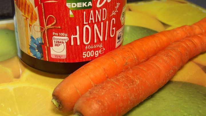 Eine Glasflasche mit Karottenhonig steht auf einem bunten Untergrund, neben zwei frischen, orangefarbenen Karotten.