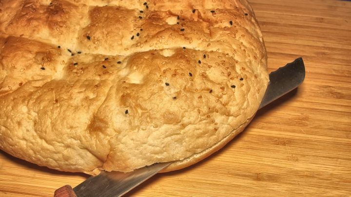 Ein Fladenbrot liegt auf einem Holzbrett, w&auml;hrend ein K&uuml;chenmesser bereit liegt, um es zu schneiden oder f&uuml;r eine Pizza-Grundlage zu verwenden.