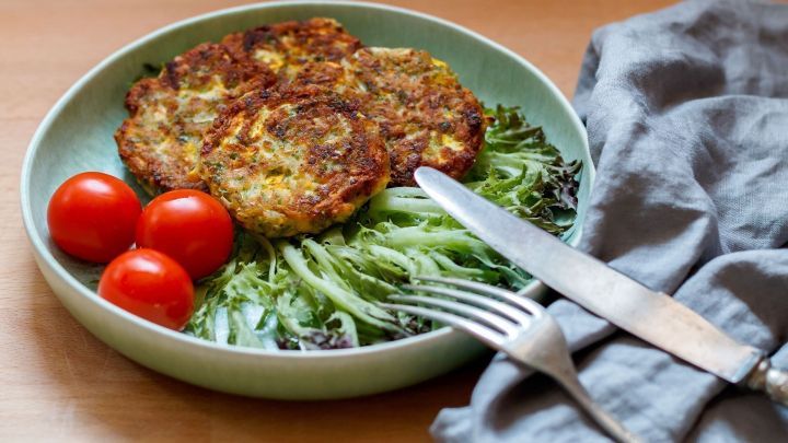 K&uuml;chle aus Zucchini liegen auf frischem Salat mit Kirschtomaten, umgeben von Besteck und einem Stoffserviette.