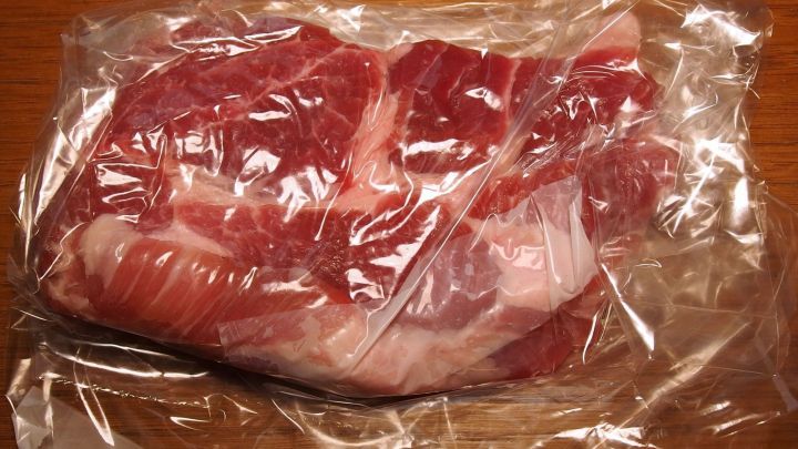 Ein rohes Schnitzel liegt in Plastikfolie auf einem Holztisch, bereit zum Klopfen ohne Spritzen.