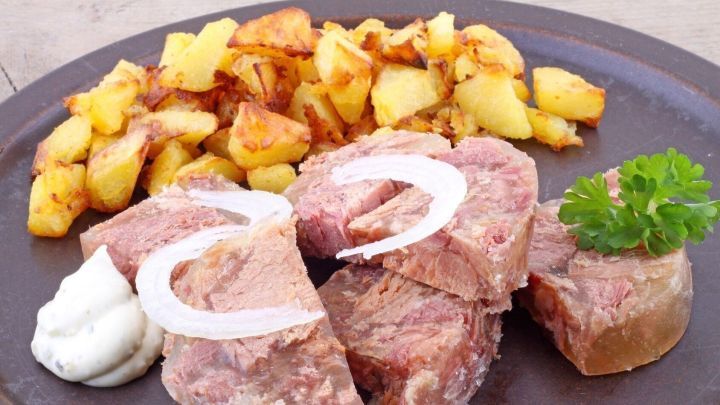 Auf einem braunen Teller liegt Sauerfleisch, dekoriert mit Zwiebelringen und Petersilie, daneben goldene Bratkartoffeln und Saucen.