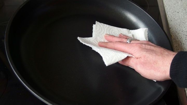 Eine Hand wischt mit K&uuml;chenpapier altes Fett aus einer Pfanne, w&auml;hrend der Hintergrund eine K&uuml;che zeigt.