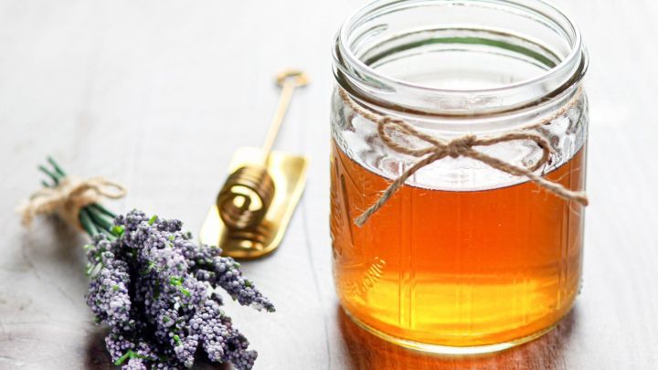 Ein Glas Honigessig steht auf einem Holztisch, umgeben von einem Lavendelstrau&szlig; und einem Honigl&ouml;ffel.