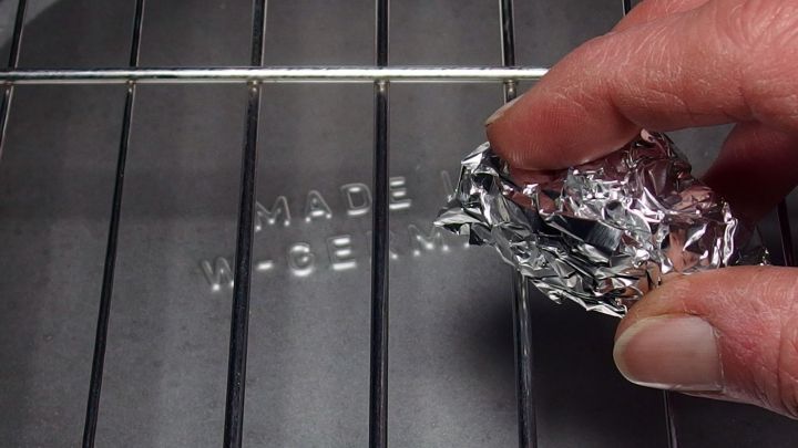 Ein Hand, die mit einer Alufolie einen Grillrost reinigt, um Schmutz zu entfernen. Der Rost ist aus Metall.