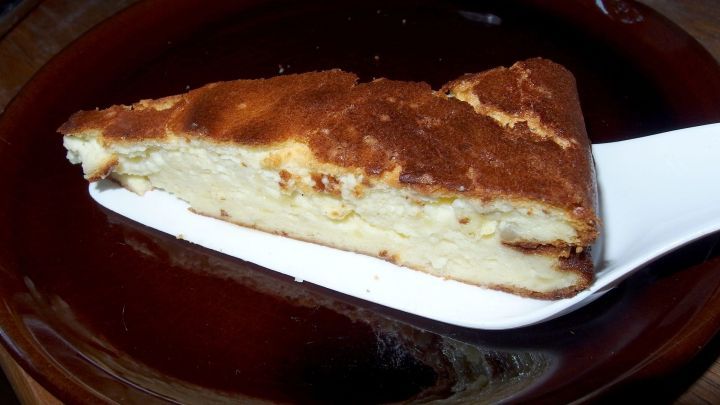 Ein St&uuml;ck K&auml;sekuchen ohne Boden liegt auf einem wei&szlig;en Teller und wird mit einem Spatel serviert. Es hat eine goldene Oberfl&auml;che.