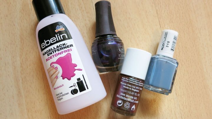 Ein Fl&auml;schchen Nagellackentferner steht neben mehreren Nagellacken auf einem Holztisch, bereit zum Verfl&uuml;ssigen.