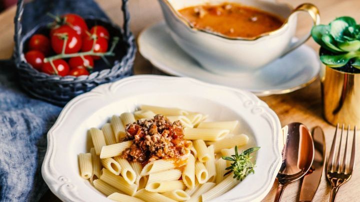 Ein Teller Nudeln mit Hackfleischsauce liegt auf einem Tisch, w&auml;hrend eine Schale Sauce daneben steht. Im Hintergrund sieht man eine kleine Obstschale.