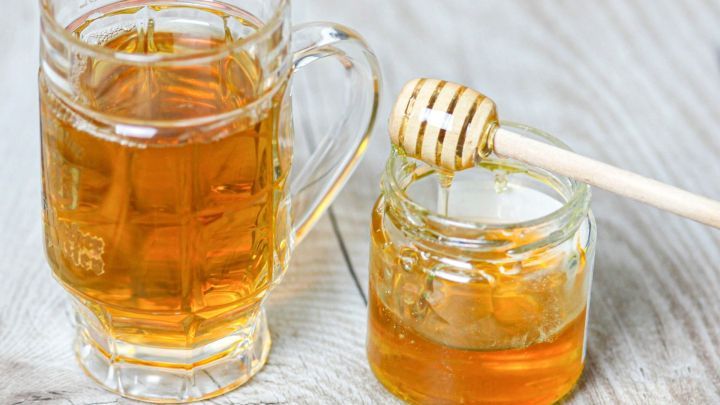 Eine Tasse mit goldenem Tee steht neben einem Glas Honig, das mit einem Holzl&ouml;ffel bedeckt ist, auf einer Holzoberfl&auml;che.