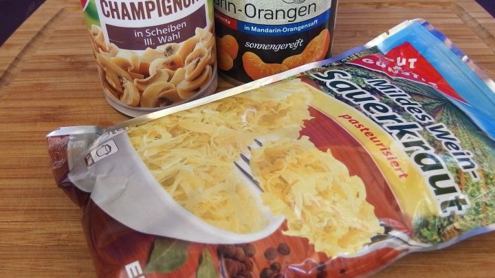 Eine Packung Sauerkraut liegt auf einem Holzbrett, neben Dosen mit Champignons und Mandarinen, bereit zum Kochen.