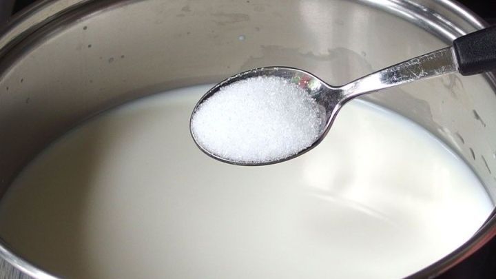 Im Topf befindet sich Milch, w&auml;hrend ein L&ouml;ffel mit Zucker dar&uuml;ber gehalten wird; die Szene zeigt eine Kochumgebung.