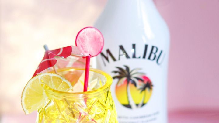 Ein gelbes Cocktailglas mit einem Malibuwasser steht vor einer Flasche Malibu auf einem pinken Hintergrund.
