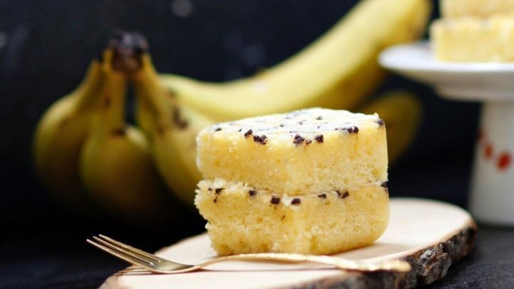 Saftiger R&uuml;hrkuchen mit einer Banane liegt auf einem Holzbrett, umgeben von frischen Bananen und einem kleinen Kuchenteller.