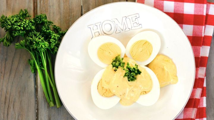 Auf einem wei&szlig;en Teller mit dem Schriftzug "HOME" liegen halbe Eier, &uuml;bergossen mit einer cremigen Senfso&szlig;e, garniert mit Fr&uuml;hlingszwiebeln. Neben dem Teller liegt ein kariertes полотенце.