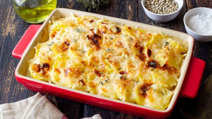 Ein goldbraun gebackenes Schnitzel-R&ouml;sti-Schmand-Gratin liegt in einer roten Auflaufform auf einem Holzbrett, umgeben von Gew&uuml;rzen.