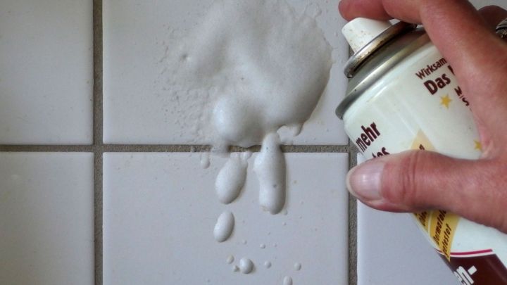 Ein Hand spr&uuml;ht Backofenspray auf einen Fliesenspiegel in der K&uuml;che, um ihn zu reinigen. Schaum spritzt an die Fliesen.