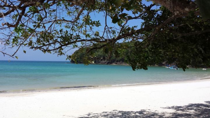 Ein wei&szlig;er Sandstrand erstreckt sich unter einem sonnigen Himmel, w&auml;hrend sanfte Wellen gegen das Ufer pl&auml;tschern.
